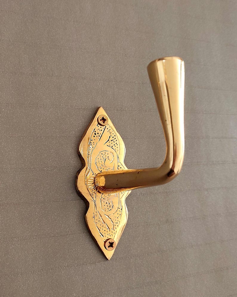 May include: A polished gold-tone metal wall hook featuring a decorative, elongated diamond-shaped backplate with intricate etched detailing. The hook is secured to the wall with two visible screws at the top and bottom of the backplate.