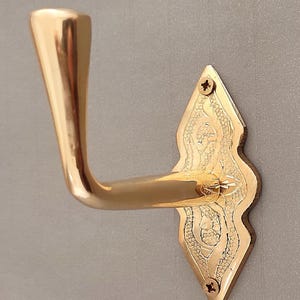 May include: A polished brass door handle with an ornate, diamond-shaped backplate. The handle curves gracefully, and the backplate features decorative etched patterns. Screws secure the backplate to the door.