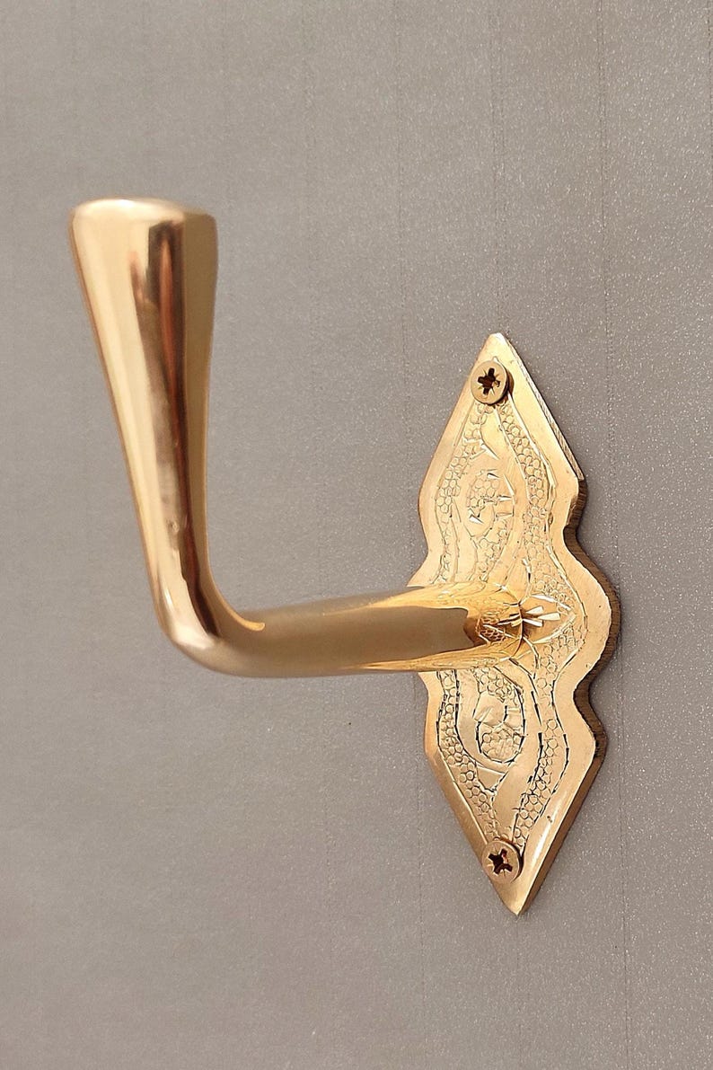 May include: A polished brass door handle with an ornate, diamond-shaped backplate. The handle curves gracefully, and the backplate features decorative etched patterns. Screws secure the backplate to the door.
