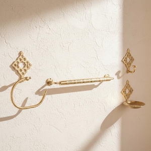 May include: A set of gold bathroom fixtures, including a towel rail, a toilet roll holder, a hook, and a small dish. The fixtures are mounted on a textured, off-white wall. Each piece has a decorative diamond-shaped element.