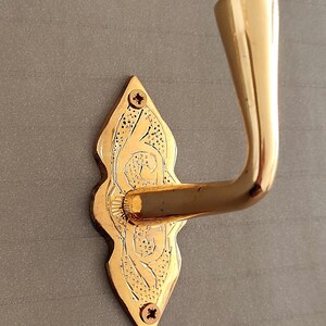 May include: A gold-coloured, ornate door hook with a decorative backplate. The backplate is shaped like a teardrop and features etched floral designs. Two screws secure the backplate to the wall. The hook curves gracefully outward.