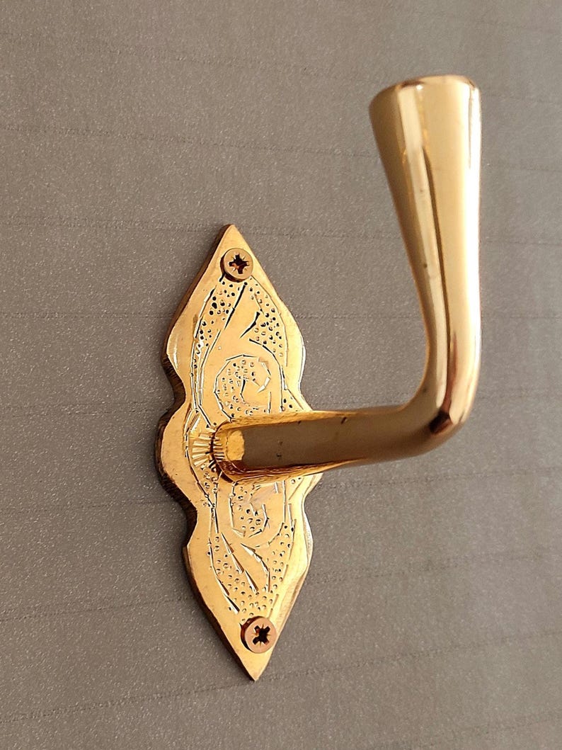 May include: A gold-coloured, ornate door hook with a decorative backplate. The backplate is shaped like a teardrop and features etched floral designs. Two screws secure the backplate to the wall. The hook curves gracefully outward.