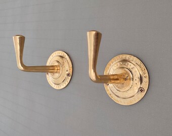 May include: Two gold-toned metal hooks with decorative circular bases. The hooks are mounted on a gray wall.