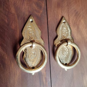 May include: Two ornate brass door handles with intricate floral designs. The handles are shaped like a teardrop with a ring pull.