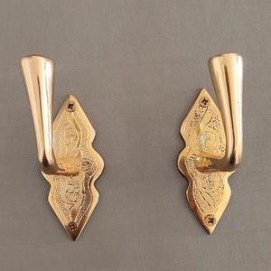 May include: Two polished brass wall hooks featuring a diamond-shaped backplate with intricate engraved floral patterns. Each hook has a smooth, curved arm extending outward and is secured to the surface with two small screws at the top and bottom.