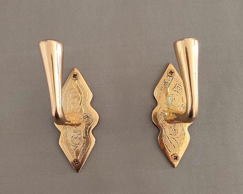 May include: Two polished brass wall hooks featuring a diamond-shaped backplate with intricate engraved floral patterns. Each hook has a smooth, curved arm extending outward and is secured to the surface with two small screws at the top and bottom.