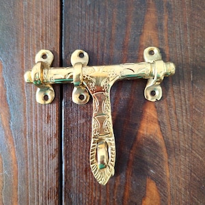 May include: A gold-toned metal door latch with an ornate design. The latch has a T-shape with a decorative handle and a locking mechanism.