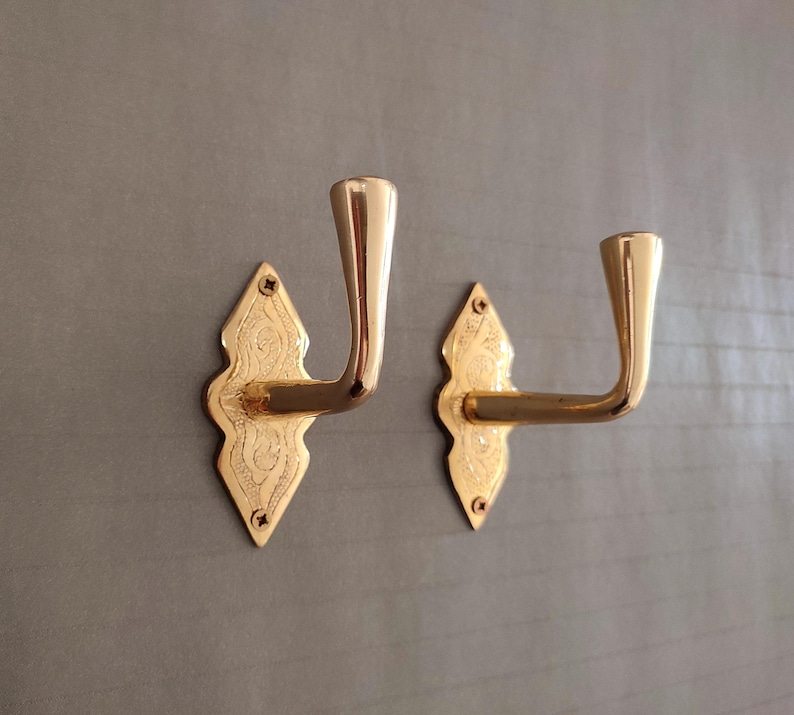 May include: A pair of gold-coloured wall hooks. Each hook features a decorative, diamond-shaped backplate with engraved details and a curved arm. The hooks are mounted on a grey surface.