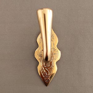 May include: A gold-toned metal wall hook featuring a smooth, curved hook arm extending from a decorative, elongated backplate. The backplate has a pointed, scalloped edge with intricate, textured engravings and a single visible screw hole at the base.