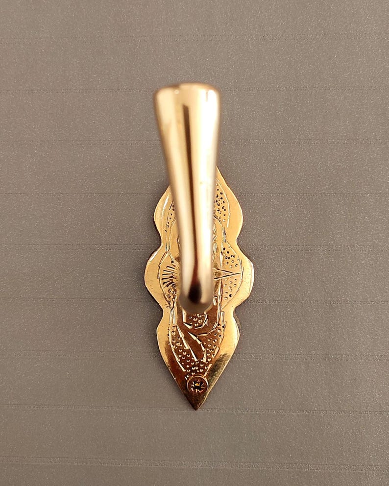 May include: A gold-toned metal wall hook featuring a smooth, curved hook arm extending from a decorative, elongated backplate. The backplate has a pointed, scalloped edge with intricate, textured engravings and a single visible screw hole at the base.
