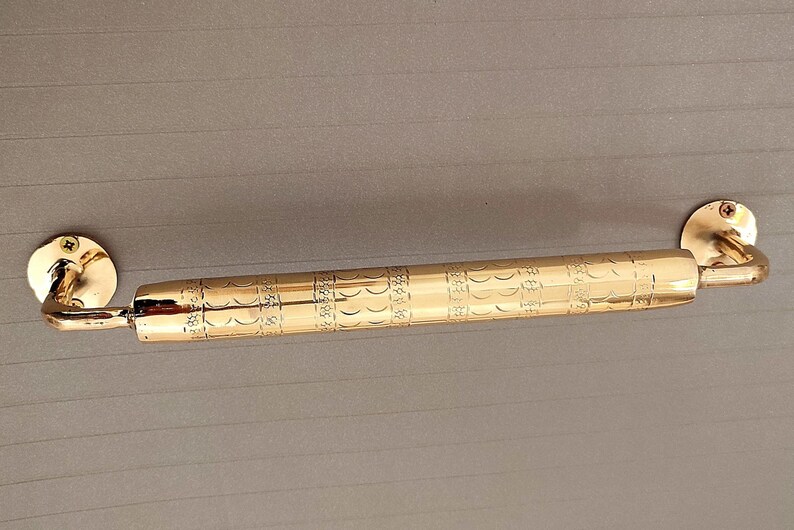 K&ouml;nnte beinhalten: Ein goldfarbener T&uuml;rgriff mit einem verzierten Design. Der Griff ist aus Metall und hat eine gebogene Form. Er ist mit Schrauben an einer Wand befestigt.