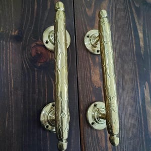 Könnte beinhalten: Zwei verzierte, goldfarbene Türgriffe mit einem blattartigen Muster. Jeder Griff ist auf einer kreisförmigen Basis mit Schraubenlöchern montiert. Die Griffe sind vor einer dunkelbraunen Holztür angebracht, was einen Kontrast in Farbe und Textur erzeugt.