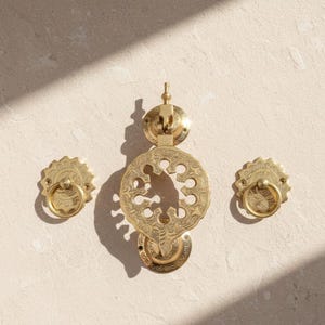 Könnte beinhalten: Ein Set aus drei Messing-Türbeschlägen. Das Mittelstück ist ein dekorativer Türklopfer mit einem verzierten, kreisförmigen Design. Zwei kleinere, passende Ringgriffe flankieren den Klopfer. Alle Teile haben eine goldene Oberfläche.