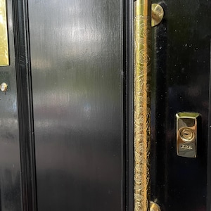 May include: A close-up of a black door with a long, ornate brass door pull. The door pull has a detailed floral pattern and is mounted vertically. An ERA door lock is also visible.