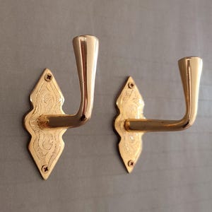 May include: Two polished brass wall hooks featuring a diamond-shaped backplate with intricate engraved detailing. Each hook has a curved, upward-facing arm and is secured to the surface with two small screws at the top and bottom points.