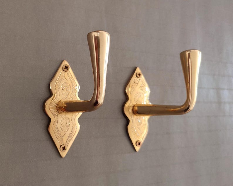 May include: Two polished brass wall hooks featuring a diamond-shaped backplate with intricate engraved detailing. Each hook has a curved, upward-facing arm and is secured to the surface with two small screws at the top and bottom points.