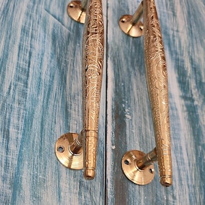 Peut inclure: Une paire de poignées de porte en laiton ornées de motifs floraux complexes. Les poignées sont longues et fines, avec une extrémité arrondie et une base plate. Les poignées sont montées sur une porte en bois.