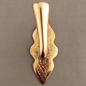 May include: A gold-coloured, ornate hook with a decorative backplate. The hook has a curved design and is attached to a detailed, leaf-shaped backplate. The backplate features intricate patterns and a small circular emblem at the bottom.
