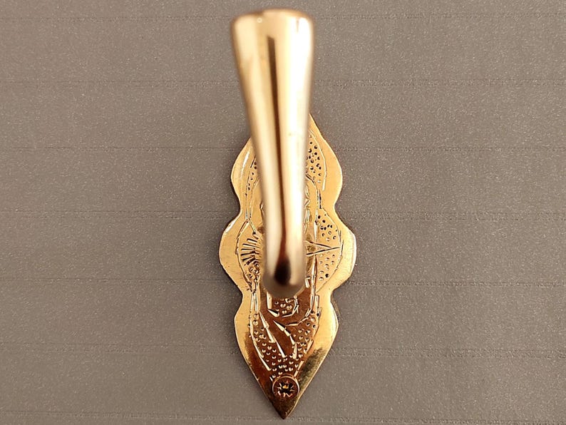 May include: A gold-coloured, ornate hook with a decorative backplate. The hook has a curved design and is attached to a detailed, leaf-shaped backplate. The backplate features intricate patterns and a small circular emblem at the bottom.