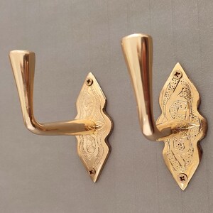 May include: Two gold-coloured wall hooks. Each hook features a curved arm and a decorative, diamond-shaped backplate with etched detailing. The hooks are mounted on a grey wall. The hooks are made of metal.