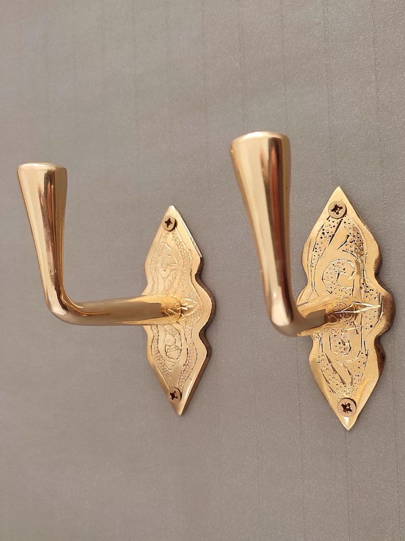 May include: Two gold-coloured wall hooks. Each hook features a curved arm and a decorative, diamond-shaped backplate with etched detailing. The hooks are mounted on a grey wall. The hooks are made of metal.
