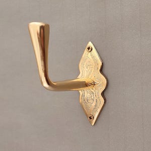May include: A polished gold-tone metal wall hook featuring a smooth, curved arm and a diamond-shaped backplate. The backplate displays an intricate, engraved floral pattern and is secured to the surface with two small, visible metal screws.