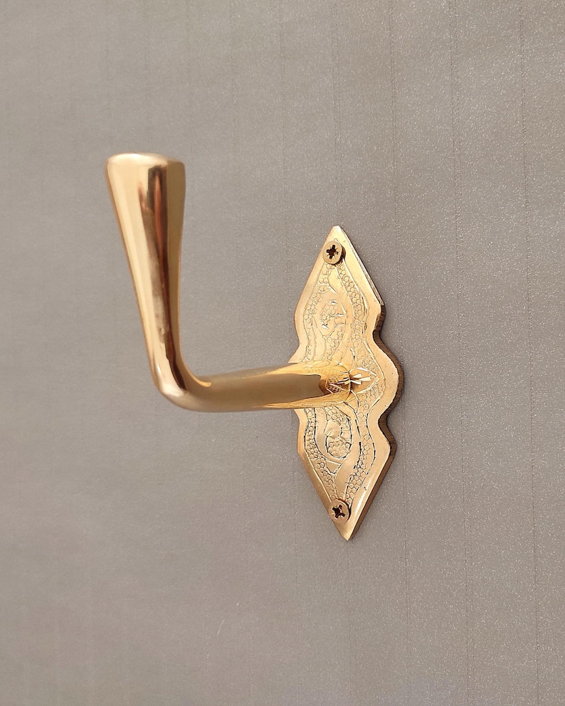 May include: A polished gold-tone metal wall hook featuring a smooth, curved arm and a diamond-shaped backplate. The backplate displays an intricate, engraved floral pattern and is secured to the surface with two small, visible metal screws.