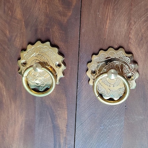 Puede incluir: Dos tiradores de armario dorados y ornamentados con tiradores de anillo. Cada tirador tiene una placa trasera decorativa y festoneada con diseños grabados de flores y volutas. Los tiradores están montados en una puerta de armario de madera marrón oscuro.