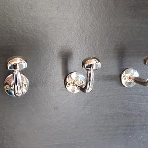 Peut inclure: Trois crochets chrom&eacute;s mont&eacute;s sur un mur gris fonc&eacute;. Chaque crochet a un sommet arrondi et un bras incurv&eacute;.