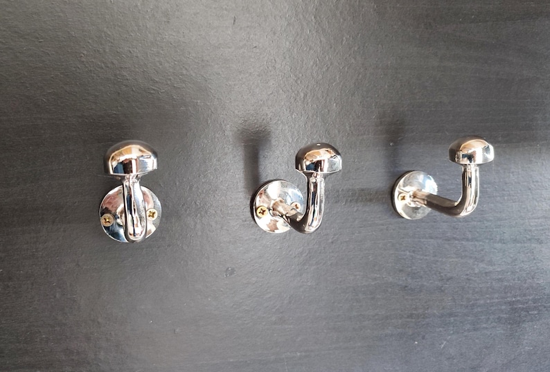 Peut inclure: Trois crochets chrom&eacute;s mont&eacute;s sur un mur gris fonc&eacute;. Chaque crochet a un sommet arrondi et un bras incurv&eacute;.