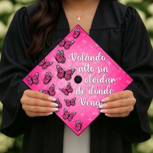 May include: A pink graduation cap decorated with pink butterflies and the Spanish text "Volando alto sin olvidar de donde vengo." The cap is held by a person wearing a black graduation gown.