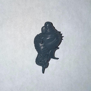 Może przedstawiać: Ciemnoszara plastikowa miniatura, prawdopodobnie do gry stołowej. Figurka ma pękaty korpus z detalami przypominającymi zbroję, kolczaste wypukłości i zakrzywiony element przypominający ostrze.