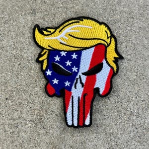 May include: An embroidered patch featuring a skull with the American flag design. The skull is wearing a yellow, stylized hairpiece.