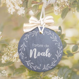 Peut inclure: Une décoration en ardoise grise avec un ruban blanc et le texte "l'arbre de Noah" en lettres blanches. La décoration est suspendue à une branche d'arbre avec des fleurs blanches.