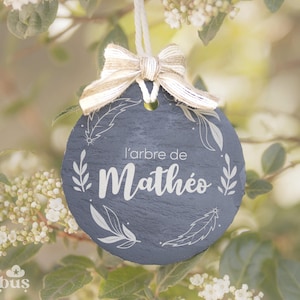 Peut inclure: Ornement rond de couleur ardoise avec l'inscription "l'arbre de Mathéo" en blanc, orné de motifs de feuilles et de plumes. Il est suspendu par une ficelle blanche et un nœud beige. L'ornement est placé sur un fond flou de verdure.