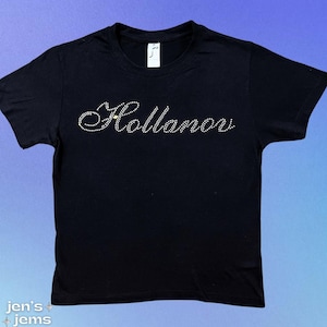 May include: Black t-shirt with the word "Hollanov" spelled out in rhinestones. The shirt has short sleeves and a crew neck. The text is in a cursive font.