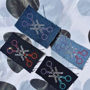 May include: Four rectangular patches featuring scissor designs. Two are denim, two are black. The scissors are decorated with rhinestones in purple, silver, red, and teal. The patches are displayed on a patterned surface.