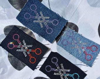 May include: Four rectangular patches with scissor designs. Two are denim, two are black. The scissors are embellished with rhinestones in various colors: purple, silver, red, and teal. The patches are arranged on a patterned surface.
