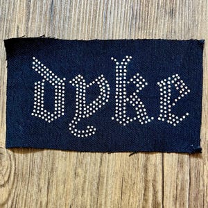May include: A rectangular, navy blue patch with the word "Duke" spelled out in white, dot-like rhinestones. The patch is set against a wooden background. The font is a gothic style.