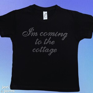 May include: Black t-shirt with the phrase "I'm coming to the cottage" in a cursive font made of small, sparkling rhinestones. The shirt has short sleeves and a crew neck.