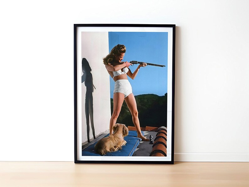 May include: A woman in a white bikini holds a rifle and aims it towards the right side of the frame. She is standing on a patio with a small dog lying on a blue cushion beside her.