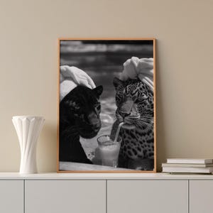 May include: Black and white photo of two animals, a black panther and a jaguar, wearing towels on their heads, sitting side by side and drinking from a glass with a straw.