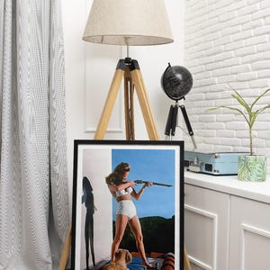 May include: A framed vintage poster featuring a woman in a white bikini holding a rifle, with a small dog sitting beside her. The poster has a blue sky and green foliage in the background.