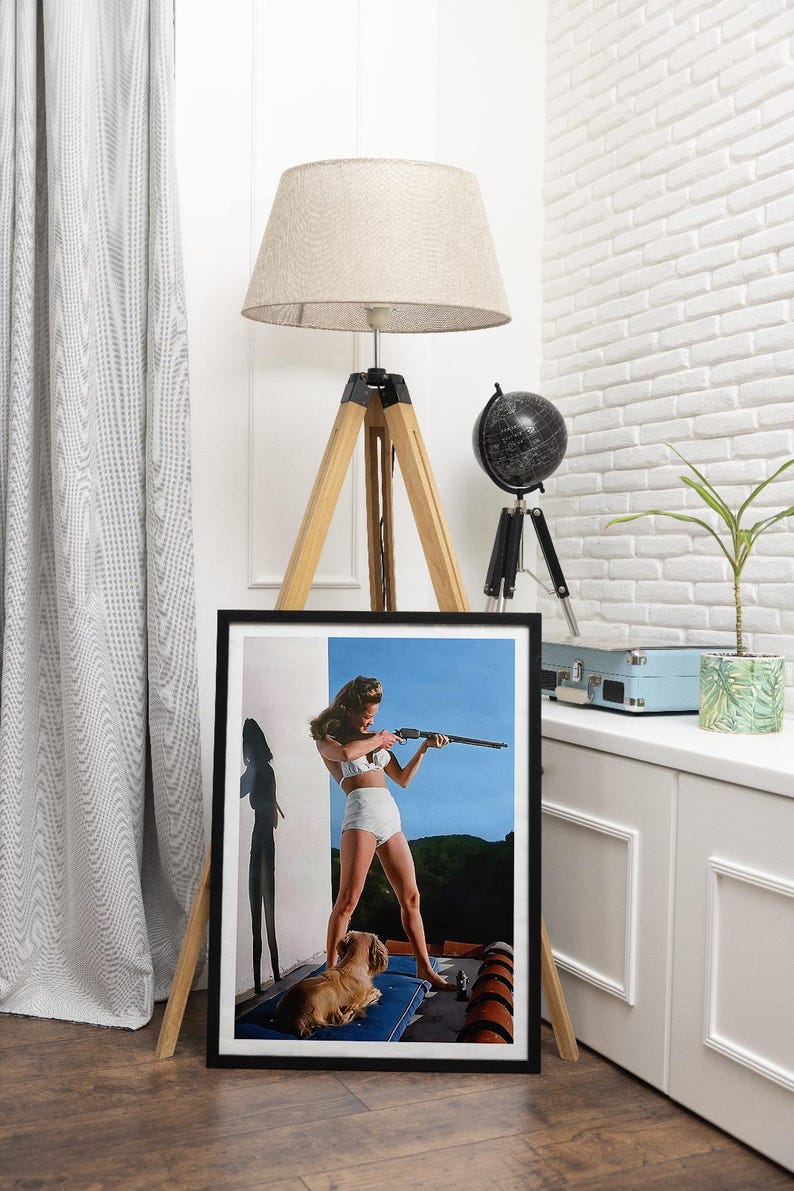 May include: A framed vintage poster featuring a woman in a white bikini holding a rifle, with a small dog sitting beside her. The poster has a blue sky and green foliage in the background.