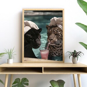 Peut inclure: Impression encadr&eacute;e repr&eacute;sentant une panth&egrave;re noire et un l&eacute;opard dans une piscine. Le l&eacute;opard boit une boisson rose avec une paille. Les deux grands f&eacute;lins portent des serviettes blanches. L'image est affich&eacute;e sur une table en bois.