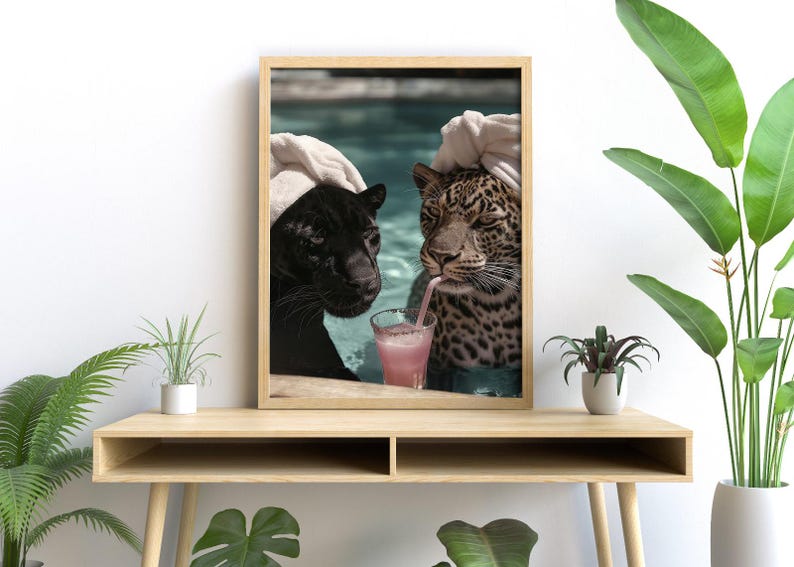 Peut inclure: Impression encadr&eacute;e repr&eacute;sentant une panth&egrave;re noire et un l&eacute;opard dans une piscine. Le l&eacute;opard boit une boisson rose avec une paille. Les deux grands f&eacute;lins portent des serviettes blanches. L'image est affich&eacute;e sur une table en bois.