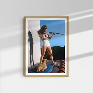 May include: A woman in a white swimsuit holds a rifle and aims it towards the right side of the frame. She is standing on a rooftop with a small dog lying at her feet. The background is a blue sky with some trees in the distance.