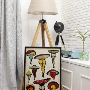 May include: A framed art print featuring whimsical mushroom illustrations with human-like legs and colorful caps. The artwork is displayed on a wooden easel in a room with a white brick wall and a floor lamp.