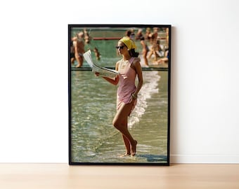 May include: A woman in a pink dress and yellow headscarf walks through the water on a beach. She is holding a newspaper in her left hand.
