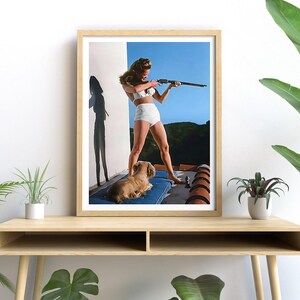 May include: A framed photograph of a woman in a white bikini holding a rifle, standing on a balcony with a small dog lying on a blue cushion.